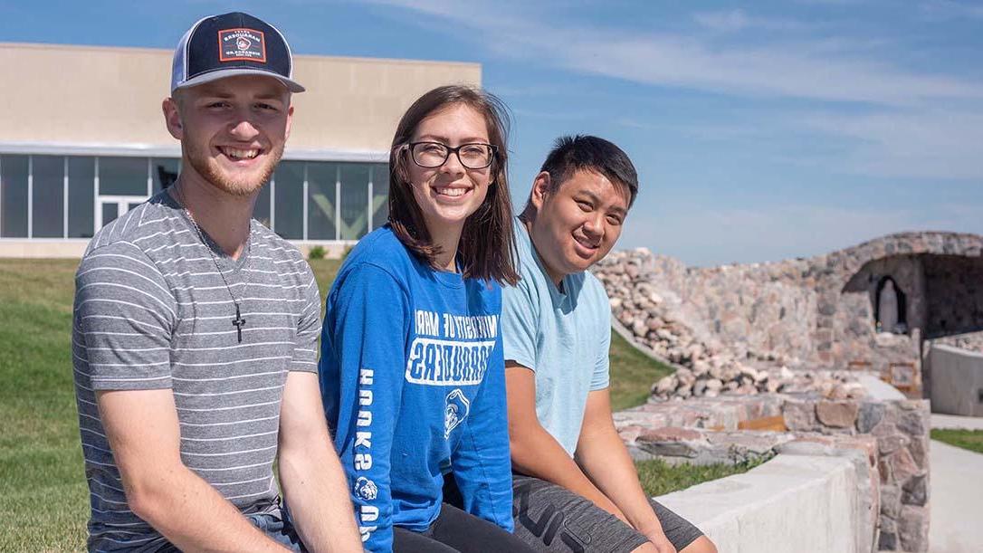 Three smiling students sitting on the edge of the rock wall leading to the grotto